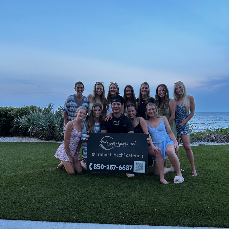 Group of women posing with a "#1 rated hibachi catering" sign on the lawn at dusk by the ocean in Clearwater Beach, Florida