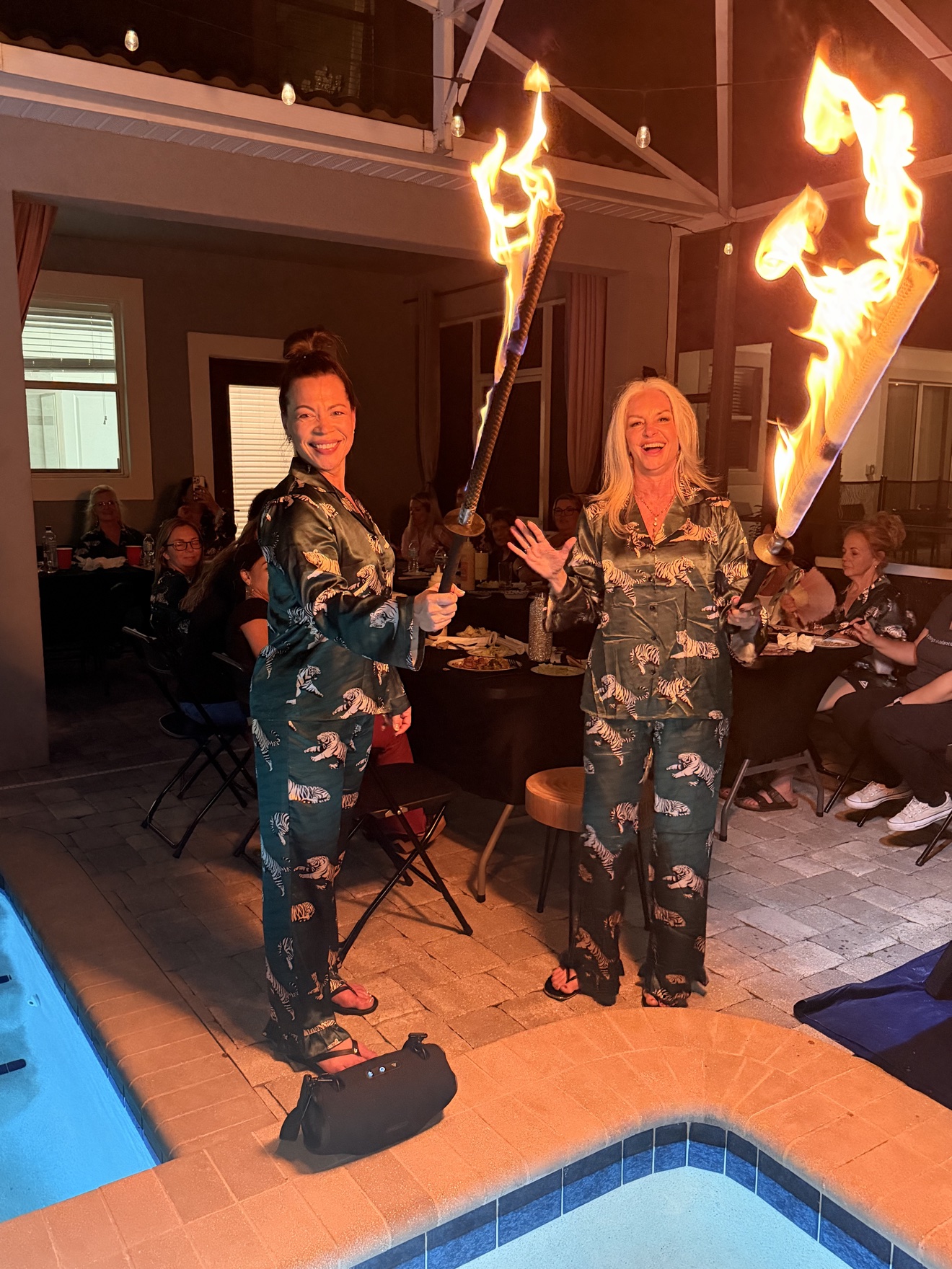 Two guests holding large fire torches by the pool at night during a private hibachi party, with other guests dining at tables behind in Aventura, Florida