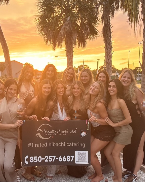Group of women posing with a "#1 rated hibachi catering" sign at sunset in Southwest Florida