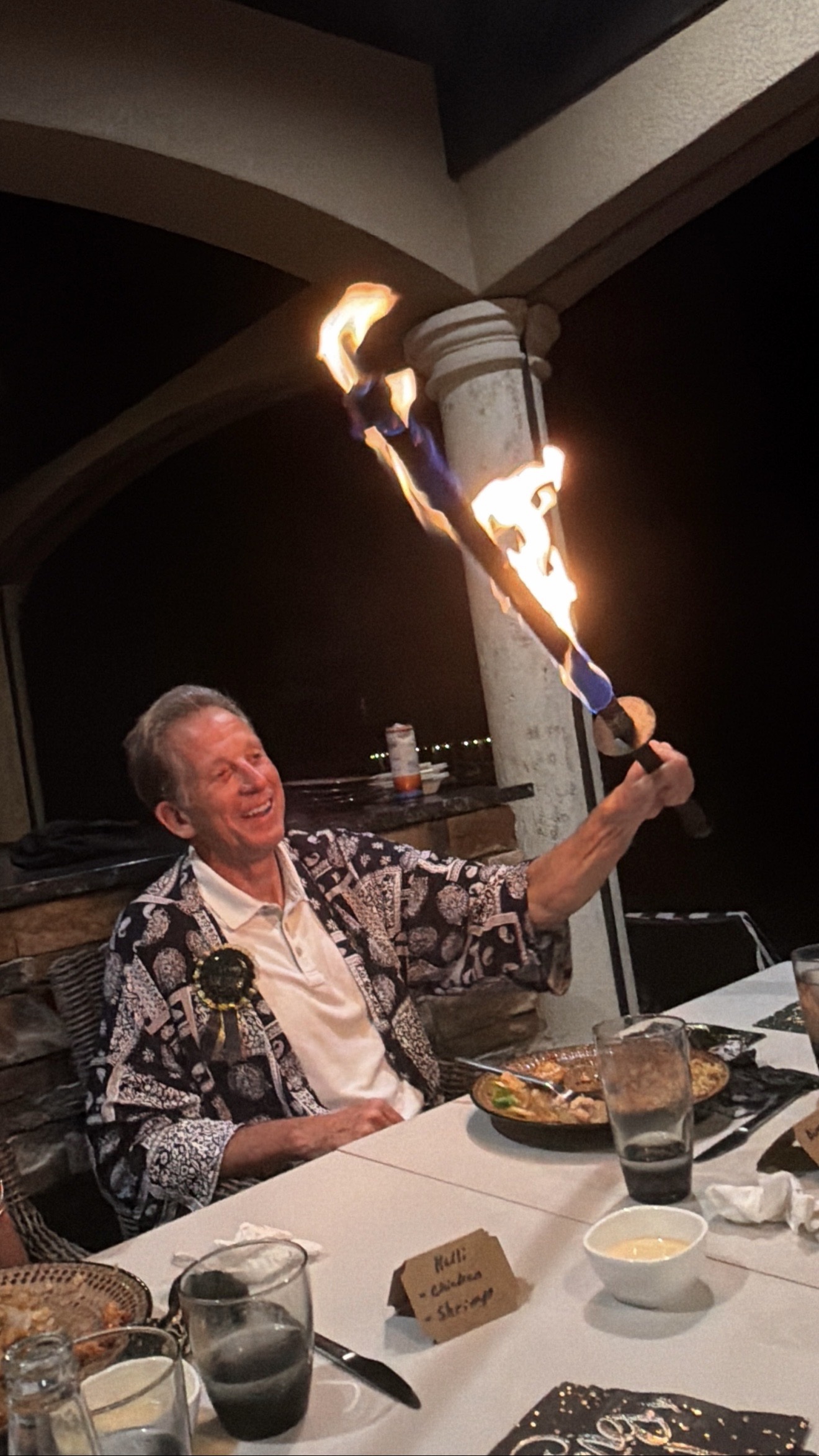 Older male guest joyfully holding up a large fire torch at a dinner table, wearing a happi coat and birthday badge, at a nighttime patio hibachi party in Hialeah, Florida