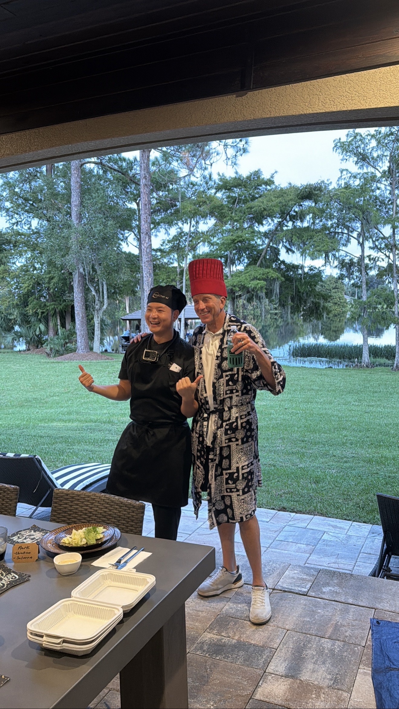 Hibachi chef posing with a male guest wearing a tall red chef hat at a lakeside patio at dusk, with lawn and lake view in Aventura, Florida