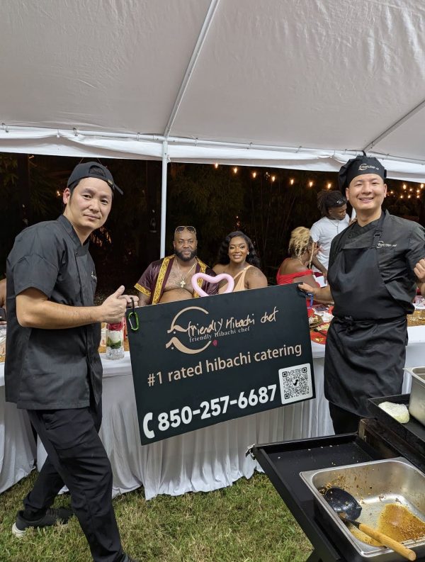 Two hibachi chefs holding a "#1 rated hibachi catering" sign at an outdoor event in Fort De Soto, Florida