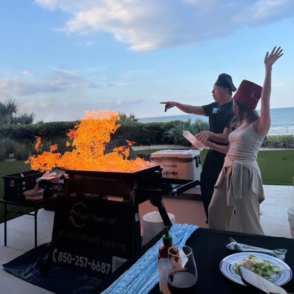 Guest cheering with arm raised as the hibachi chef creates a huge flame burst by the ocean at dusk