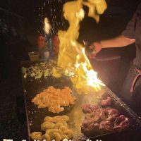Dramatic hibachi fire show with flames and sparks erupting over shrimp and steak on the grill at night